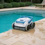 Robot de piscine sans fil posé au bord d'une piscine rectangulaire extérieure avec eau turquoise et carrelage en pierre claire